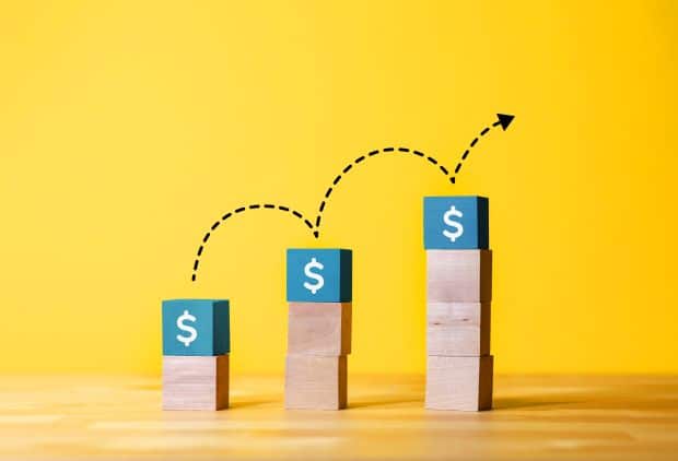 A stack of wooden blocks on a yellow background with an arrow pointing to a dollar sign. The blocks represent the marketplace, showcasing effective marketing management by My Amazon Guy.